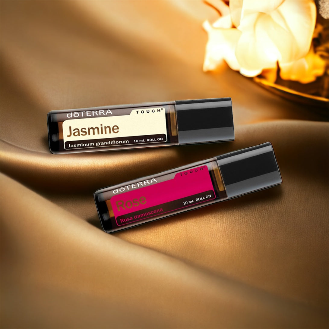 Two bottles of essential oil labeled doTerra Rose Touch and doTerra Jasmine Touch, on a gold silken sheet with an illuminated white rose above.