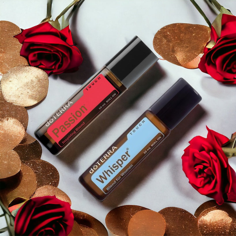 Two rollerball bottles of doTerra essential oils, one labeled 'Passion' in red and the other 'Whisper' in a pale color, presented on a white surface with gold disks and red roses.