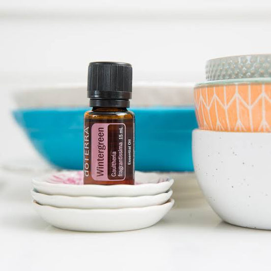 doTERRA Wintergreen essential oil bottle on a white surface with decorative bowls in the background.