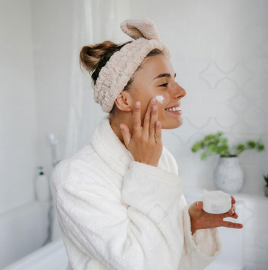 Woman applying cream to her face wearing a white robe and headband in a bathroom setting.