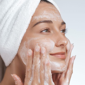 Woman applying cream to her face with a towel on her head against a white background
