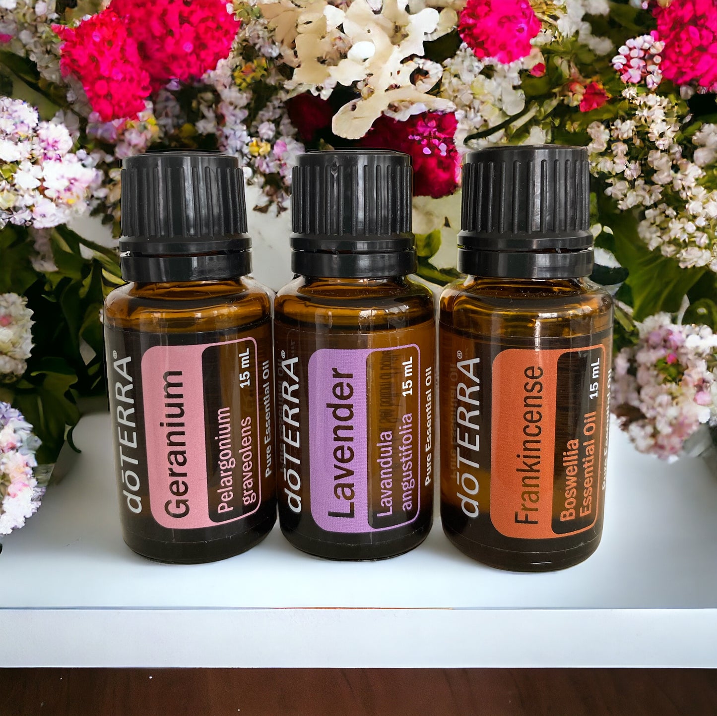 Three doTerra essential oil bottles, labeled Frankincense, Geranium, and Lavender, arranged side by side with a vase of flowers in the background.