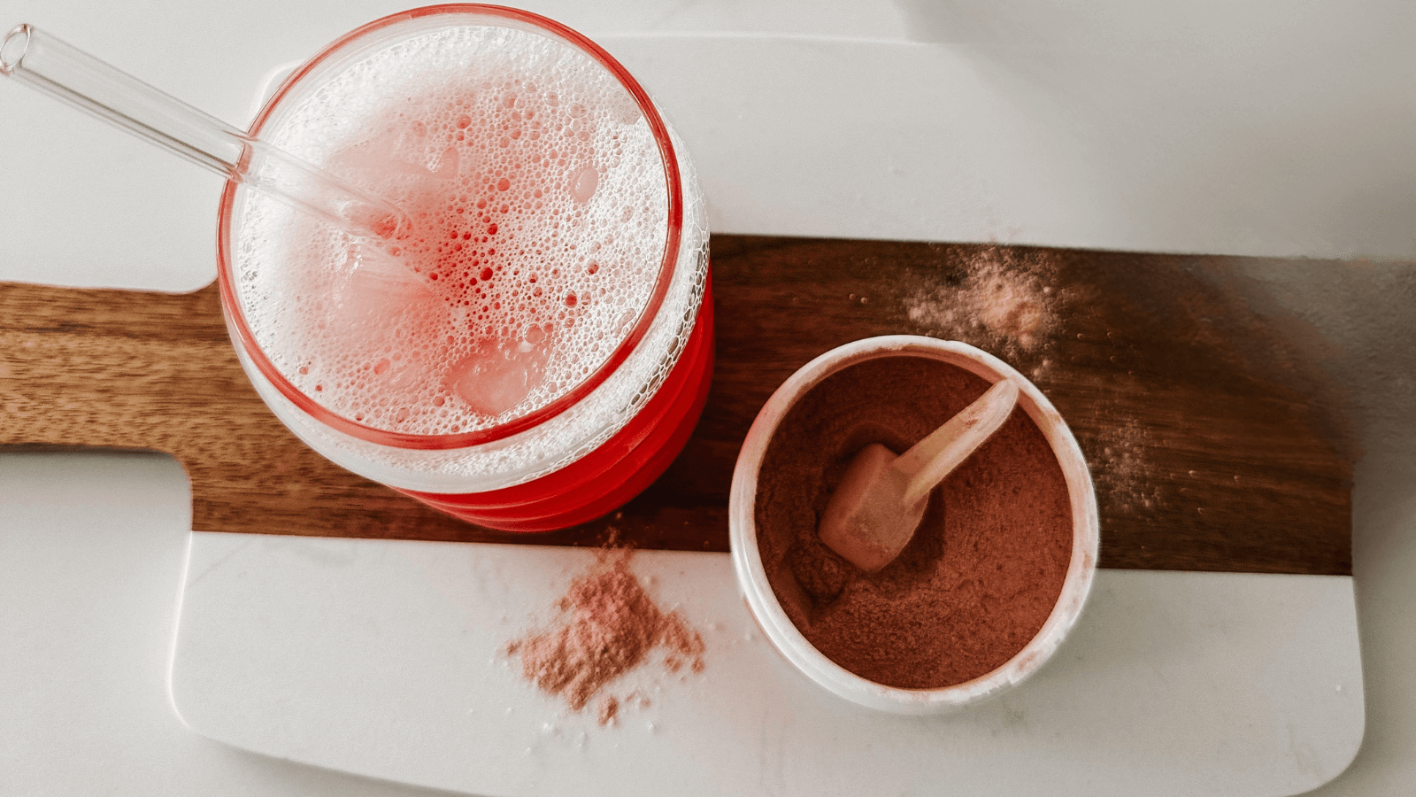 Red glass with Amare Happy juice a frothy beverage on a wooden board next to a bowl of Amare edge powder in a white tub.