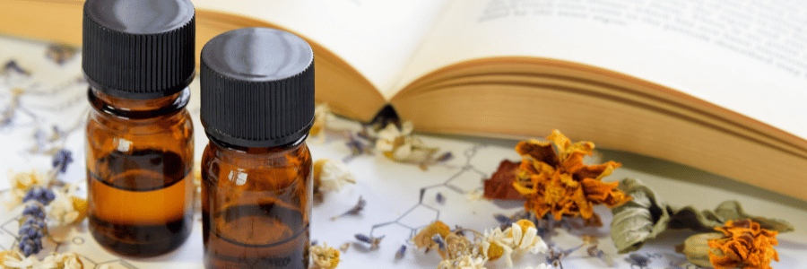 Two small amber glass bottles with black caps on a white surface with dried flowers and an open book.