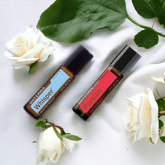 Two rollerball bottles of doTerra essential oils, one labeled 'Passion' in red and the other 'Whisper' in a pale color, presented on a white surface with green leaves and white roses.