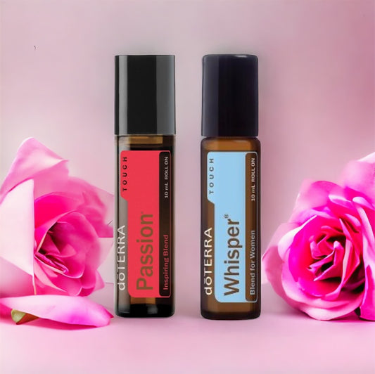 Two rollerball bottles of doTerra essential oils, one labeled 'Passion' in red and the other 'Whisper' in a pale color, presented on a pink surface with dark pink roses.