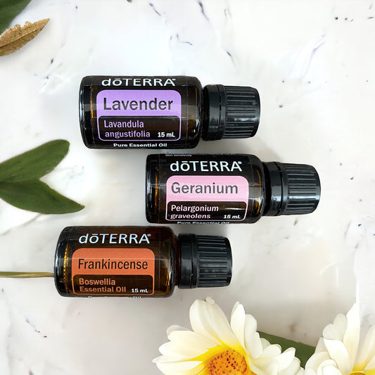 Three doTerra essential oil bottles, labeled Frankincense, Geranium, and Lavender, scattered on a white bench top with white flowers next to them.