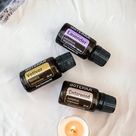 doterra vetiver, doterra lavender and doterra cedarwood essential oils laying scattered on a white cloth with a candle next to them.