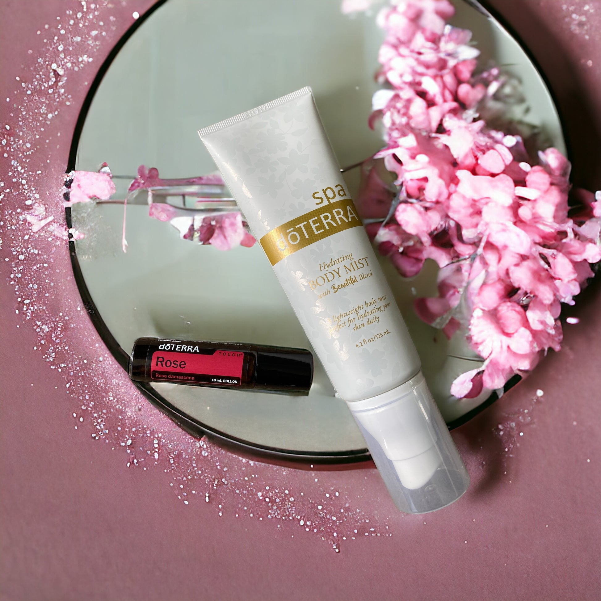doterra hydrating mist and doterra rose touch laying on a circular mirror against a pink background with pipnk flowers.